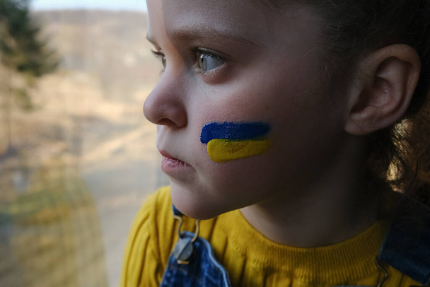 Sign of Ukrainian flag on child's cheek. Ukrainian geopolitics globe crisis. Unhappy little girl kid sitting alone on windowsill at home. Sadness longing hope. No war. Anxiety and anticipation - Fotó, kép