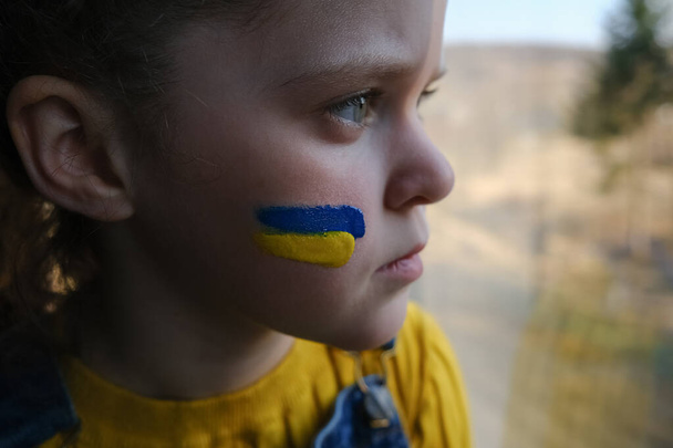 Side view of little child girl with flag of ukraine on face, kid sitting on windowsill. Peace, no war, help, stop russian aggression. Evacuation of civilians. Freedom to Ukraine, Crisis concept - Valokuva, kuva