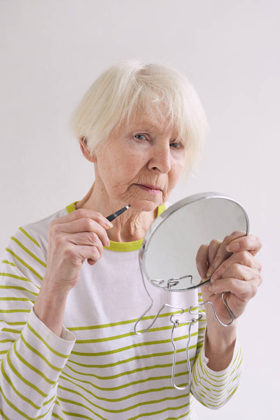 Valkoihoinen vanhempi nainen, jolla on kulmakarvat ja pieni peili nyppimässä partaansa. Kauneus, ikämuutokset, hoitokonsepti - Valokuva, kuva