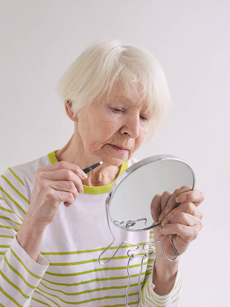 Caucasian senior woman with eyebrow tweezers and small mirror plucking her beard. Beauty, age changes, care concept - Photo, Image