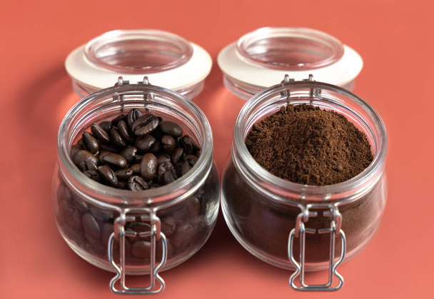 Ψητά φασόλια καφέ και αλεσμένο καφέ σε γυάλινα βάζα κοντά σε ροζ φόντο. Αντιπροσωπεύουν πρωινό, ενέργεια, φρεσκάδα ή άρωμα - Φωτογραφία, εικόνα