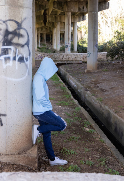 Κουκουλοφόρος έφηβος σε ανοιχτό μπλε υπερμεγέθη κουκούλα παραμένει κάτω από τη γέφυρα. Έφηβη μένει έξω κοντά σε κολώνες γέφυρας. Έννοια hipster και εφηβείας - Φωτογραφία, εικόνα