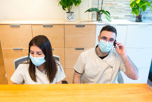 Dos recepcionistas con mascarilla facial mientras trabajan en la recepción de un centro de fisioterapia. - Foto, Imagen