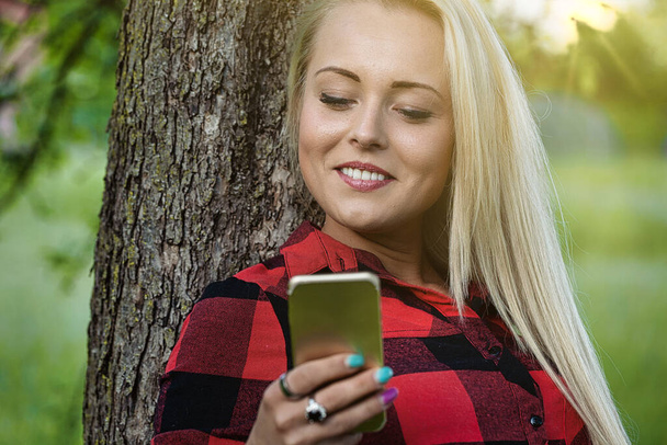 Jonge vrouw leunend tegen een boomstam in een park met een mobiele telefoon in haar handen glimlachend vrolijk verlicht door de warme gloed van de zon door bomen - Foto, afbeelding