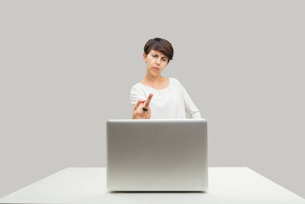 Angry ordinary young woman gestures toward her laptop screen, erects middle finger toward what she disagrees with: injustice, evil - 写真・画像