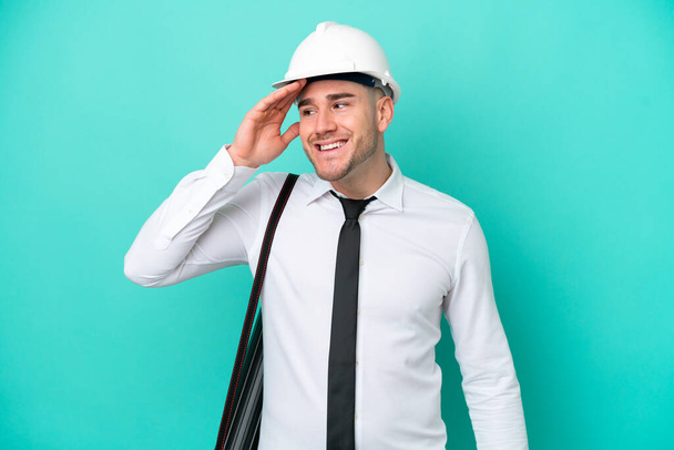Giovane architetto uomo caucasico con casco e tenendo cianografie isolate su sfondo blu sorridente molto - Foto, immagini