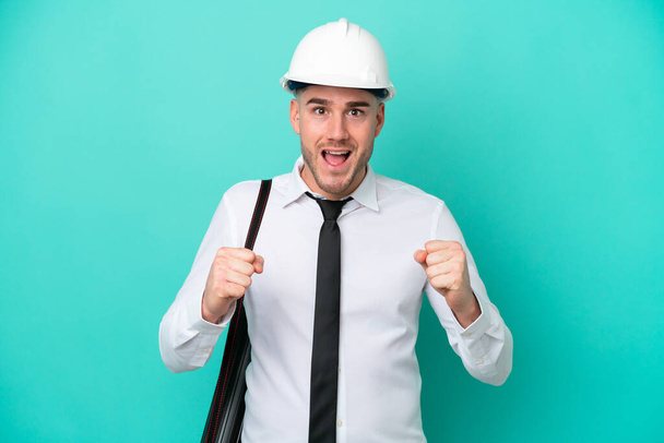 Young architect caucasian man with helmet and holding blueprints isolated on blue background celebrating a victory in winner position - Photo, Image