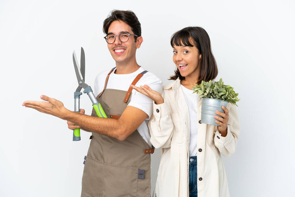 Young mixed race gardeners holding a plant and pruning shears isolated on white background extending hands to the side for inviting to come - Foto, Imagem