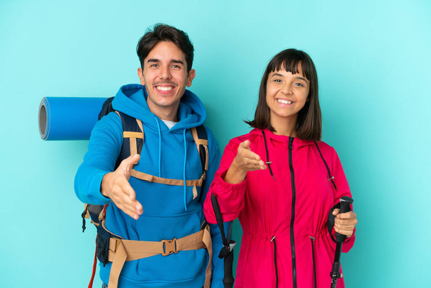 Young mountaineers couple isolated on blue background shaking hands for closing a good deal - Fotoğraf, Görsel