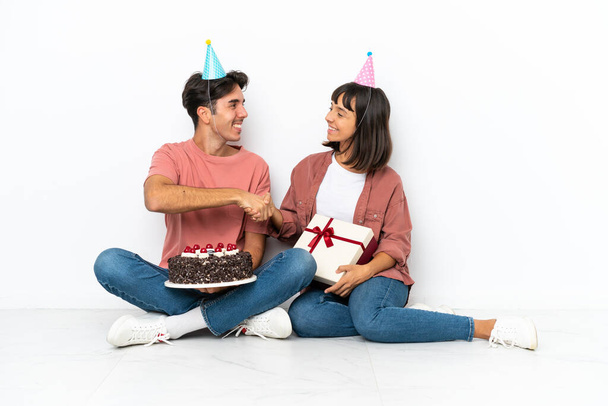 Giovane coppia di razza mista che celebra un compleanno seduto sul pavimento isolato su sfondo bianco stretta di mano dopo un buon affare - Foto, immagini