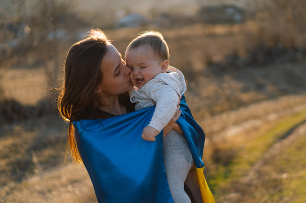 Женщина обнимает своего маленького сына, завернутого в желто-синий флаг Украины на открытом воздухе. - Фото, изображение