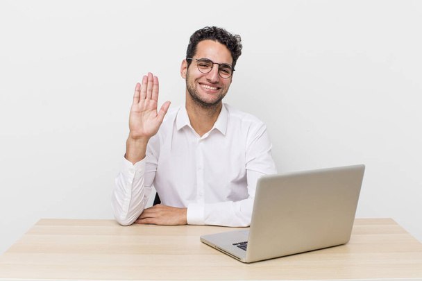 hispanic handsome man smiling happily, waving hand, welcoming and greeting you. businessman and desk concept - Foto, Bild