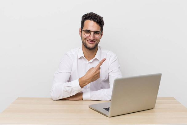 hispanic handsome man smiling cheerfully, feeling happy and pointing to the side. businessman and desk concept - Фото, изображение