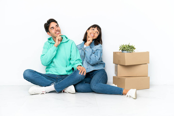 Young couple making a move while picking up a box full of things sitting on the floor isolated on white background thinking an idea while looking up - Photo, Image