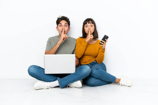 Młoda para z laptopem i telefonem siedząca na podłodze, pokazująca znak milczenia, wkładająca palec do ust. - Zdjęcie, obraz