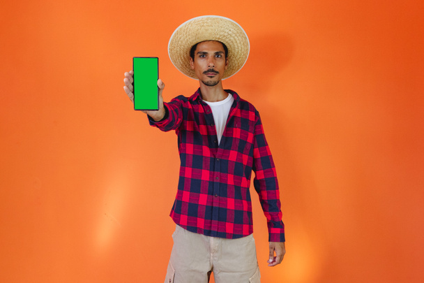 Junina Party Kıyafetli Siyah Adam elinde bir tablet ya da portakal arkaplanda izole edilmiş bir cep telefonu tutuyor. Festa Junina için geleneksel kıyafetler giyen genç adam - Brezilya Haziran Festivali. - Fotoğraf, Görsel