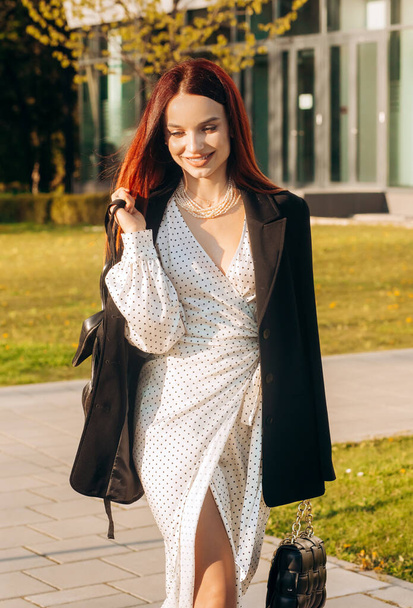 Stylish red-haired woman in a white dress and black jacket walking down the street near the office building - Zdjęcie, obraz