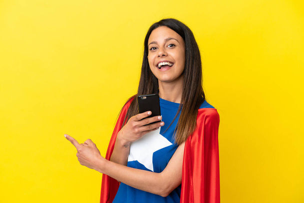 Super Hero woman isolated on yellow background using mobile phone and pointing back - Photo, image