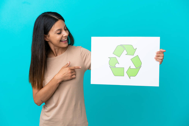 Mujer joven sobre fondo aislado sosteniendo un cartel con icono de reciclaje y señalándolo - Foto, imagen