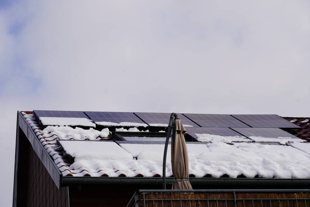 Solarmodule auf dem Dach eines Wohnhauses im Winter. Die oberen Module sind bereits schneefrei, die unteren Module noch bedeckt. Der Himmel ist fast komplett grau, kaum Strom lässt sich erzeugen. - Foto, Bild