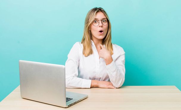 pretty blonde woman looking shocked and surprised with mouth wide open, pointing to self. business and desk concept - Foto, imagen
