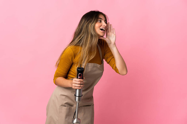 Giovane chef donna utilizzando frullatore a mano isolato su sfondo rosa gridando con la bocca spalancata al lato - Foto, immagini