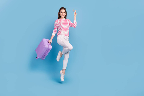 Full size portrait of attractive cheerful lady hold suitcase hand demonstrate v-sign isolated on blue color background - 写真・画像