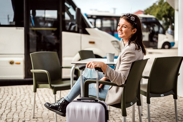 Молодой путешественник с багажом и защитной маской сидит в кафе и ждет приезда автобуса - Фото, изображение