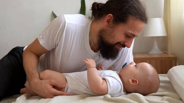 Authentic Young Bearded Man Holding Newborn Baby. Dad And Child Son On Bed. Close-up Portrait of Smiling Family With Infant On Hands. Happy Marriage Couple On Background. Childhood, Parenthood Concept - Photo, Image