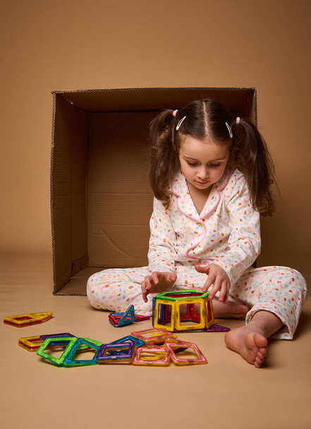 Euroopan pieni tyttö kaksi poninhäntää pyjama leikkii värikäs magneettinen rakentaja lelu beige tausta kopio mainostilaa - Valokuva, kuva