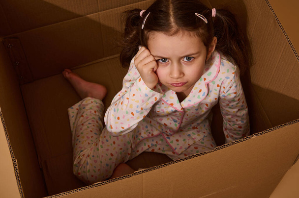 Студия снимает красивую расстроенную маленькую девочку, которая смотрит в камеру в разочаровании, находясь внутри картонной коробки. Эмоциональный студийный портрет на бежевом фоне с копией рекламного пространства - Фото, изображение