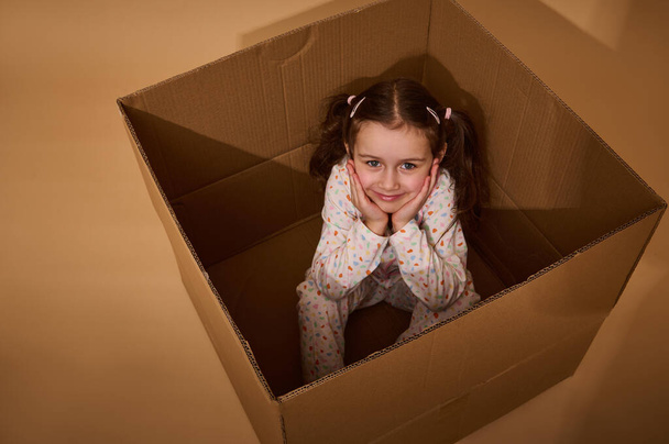 Vista aérea de una encantadora niña caucásica en pijama con puntos coloridos sentados dentro de una caja de cartón y mirando a la cámara, aislados sobre fondo beige con espacio para copiar anuncios - Foto, Imagen