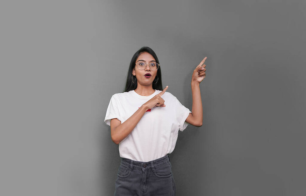 молода азіатська індійська жінка в білій футболці на сірому фоні вказуючи пальцем праворуч від себе
. - Фото, зображення