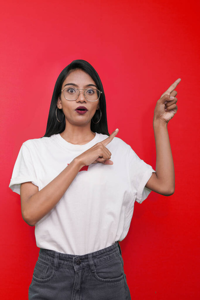 nuori aasialainen intialainen nainen yllään valkoinen t-paita vastaan punainen tausta osoittaa sormella oikealla puolella itseään. - Valokuva, kuva