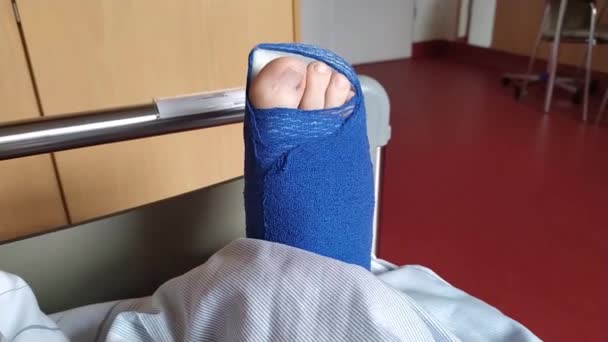 Hombre europeo con ruptura del tendón de Aquiles acostado en la cama del hospital después de la operación con zapato especial y sala de muletas para la rehabilitación médica después de la emergencia - Metraje, vídeo