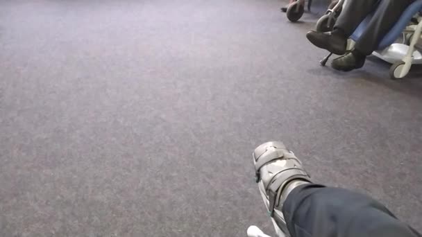 Hombre europeo bruja Aquiles ruptura tendón sentado en la sala de espera del hospital después de la operación con zapato especial y muletas en la sala de espera vacía para la rehabilitación médica después de la emergencia - Metraje, vídeo