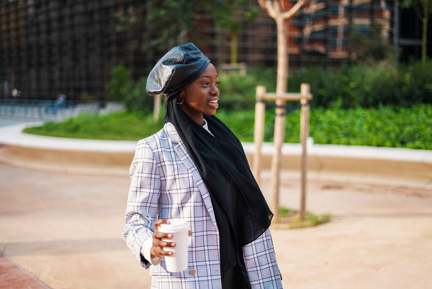 Mujer negra sonriente en bufanda y con café en el parque de la ciudad - Foto, imagen
