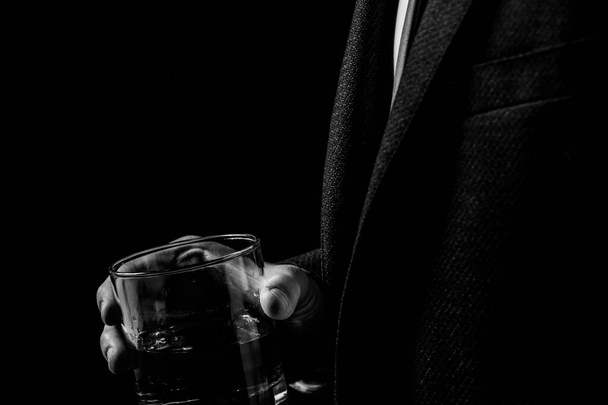 Een glas whisky in de handen van een man in een pak in het donker, een man drinkt, zwart-wit foto - Foto, afbeelding