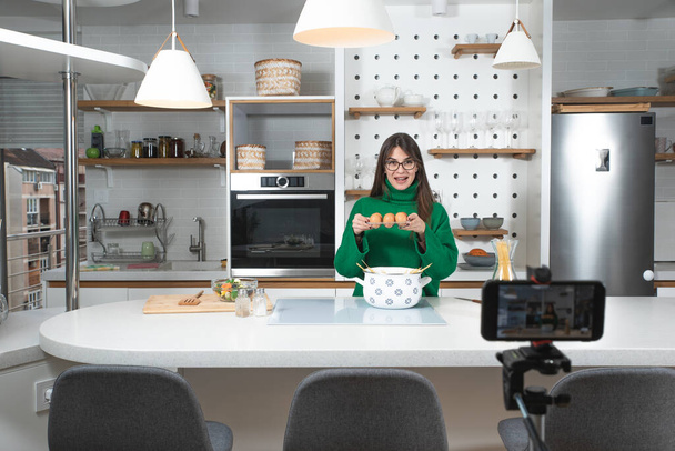 Young vlogger woman vlogging about cooking at her apartment domestic kitchen cook and making video vlog with smartphone as online content creator for internet. Female influencer making a meal. - Фото, зображення