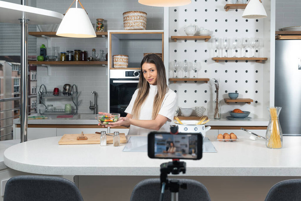 Young vlogger woman vlogging about cooking at her apartment domestic kitchen cook and making video vlog with smartphone as online content creator for internet. Female influencer making a meal. - Фото, зображення