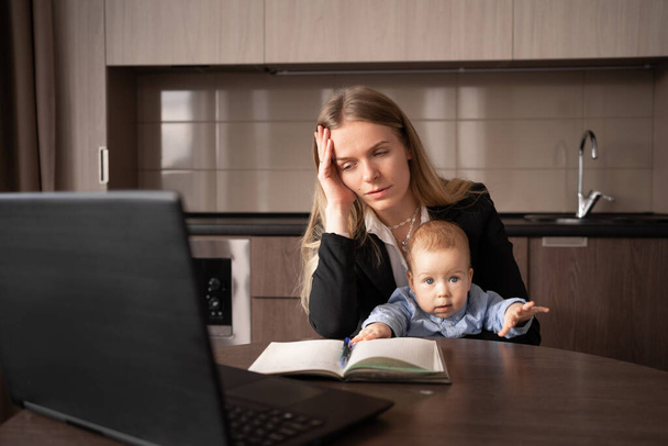 Doğum izni stresi. Kollarında bebek olan genç bir kadın ev bilgisayarında çalışırken baş ağrısından muzdarip, hasta bir anne aynı anda birden fazla iş yapamıyor. - Fotoğraf, Görsel
