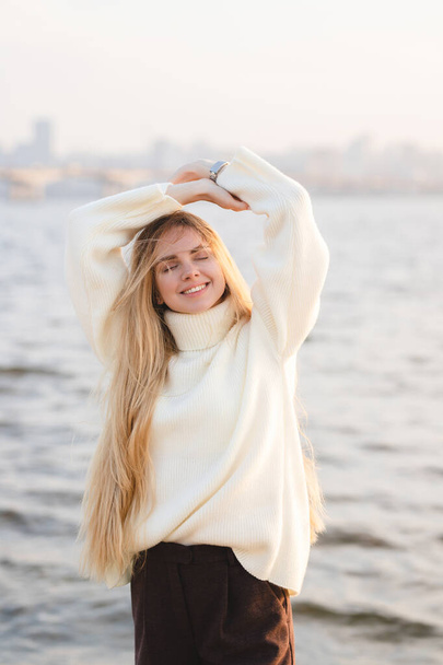 Długowłosa blondynka na brzegu rzeki. Szczęśliwa młoda dziewczyna relaksuje się w jesienny dzień - Zdjęcie, obraz