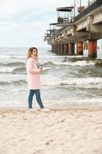 海岸を歩いてる女の子。ブルネットはコーヒーを飲む。ピンクのコートの女の子。海 - 写真・画像