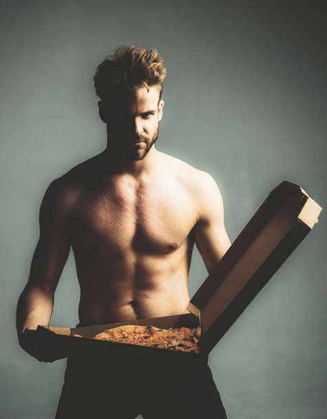 Strong brutal bearded man holding an italian tasty pizza isolated at gray background. Sexy pizza deliver with naked muscular torso. Handsome hungry man with food fetish. - Foto, Imagen