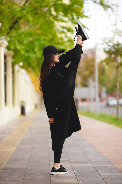 Κορίτσι γυμνάστρια με σηκωμένο πόδι πάνω από το κεφάλι της σε δρόμο της πόλης. Ένα ελκυστικό κορίτσι με παλτό και καπέλο του μπέιζμπολ ασχολείται με γυμναστική στο δρόμο. - Φωτογραφία, εικόνα