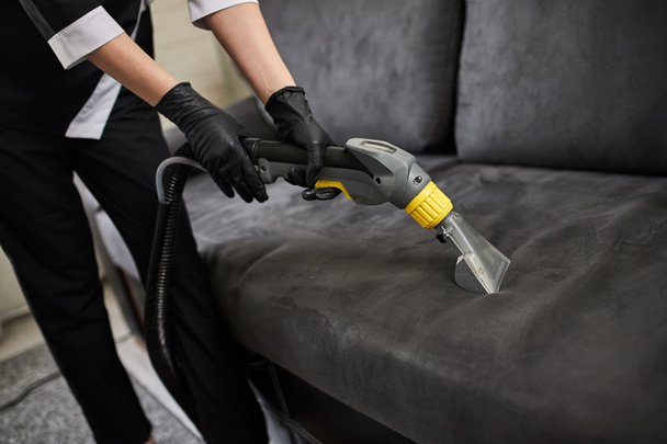 Pracovník úklidové společnosti odstraňuje nečistoty z nábytku v bytě s profesionálním vybavením. Žena hospodyně čištění ramen pohovka s prací vysavač zblízka - Fotografie, Obrázek