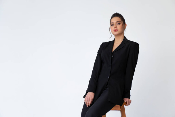 Young fashionable business woman sitting high bar wooden chair dressed black office suit isolated in white background. Caucasian female business person pensive looking at camera - Фото, изображение