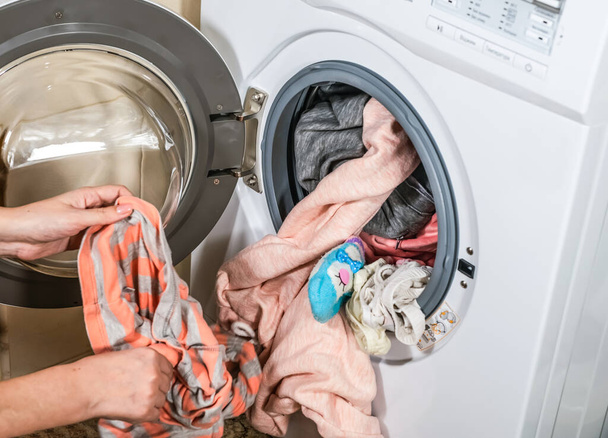 Het proces van het lossen van gewassen kleurrijke kleren uit de wasmachine in de badkamer - Foto, afbeelding