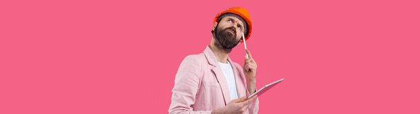 Portrait of young construction engineer wear orange hard hat, in a pink jacket standing on red studio background. A man with a tablet in his hands. - Фото, зображення