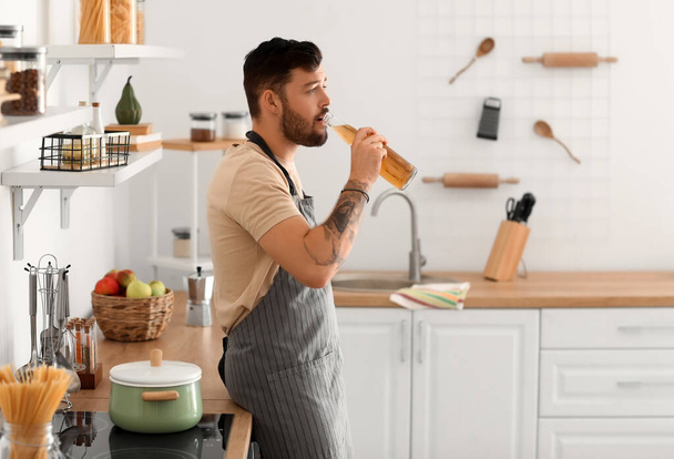 Молодой бородатый мужчина пьет пиво на кухне - Фото, изображение